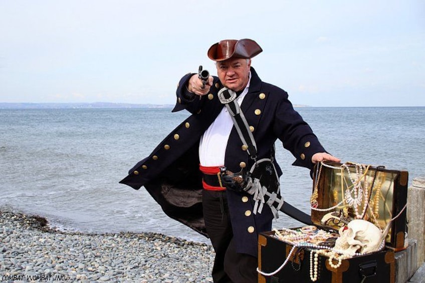 A person dressed as a pirate with a sword, pointing a gun, and holding a treasure chest on a beach. 1