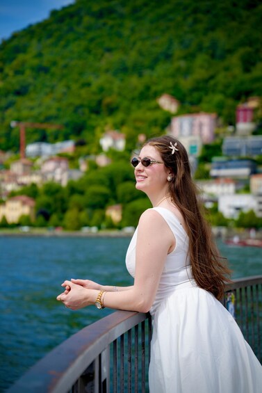 A person in a white dress leaning on a railing by a body of water with a hillside in the background. 1