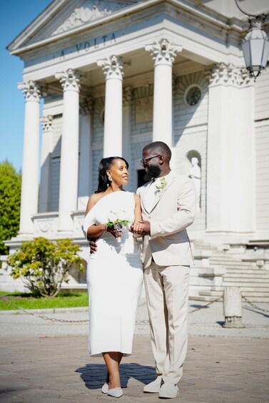 A couple in wedding attire standing in front of a building with columns and the inscription 'A VOLTA.' 1