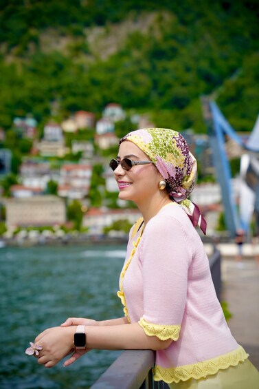 A person wearing a pink blouse and a patterned headscarf, standing by a body of water with a bridge and buildings in the background. 5