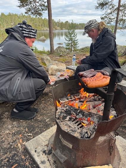 Two people cooking hot dogs over an open fire in a forested area. 2