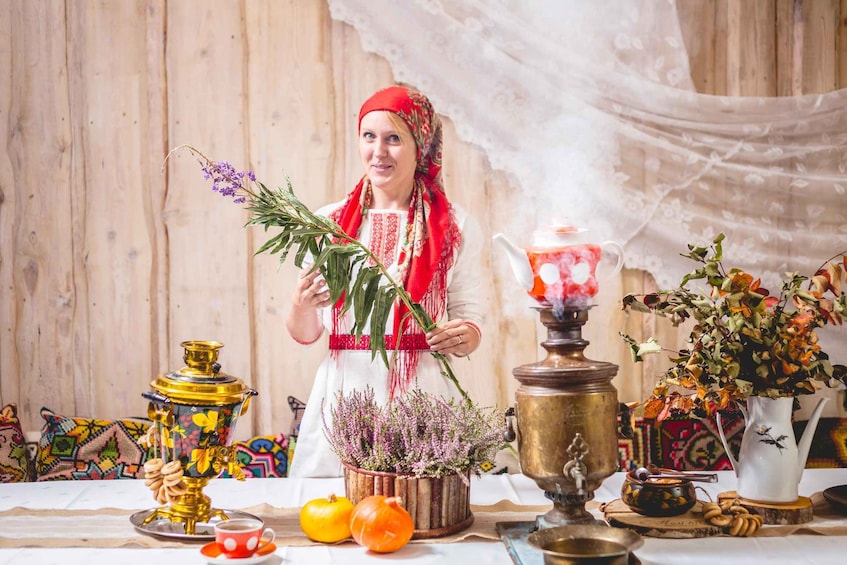 A person in traditional attire holding a plant, with samovars and a teapot on a table. 1