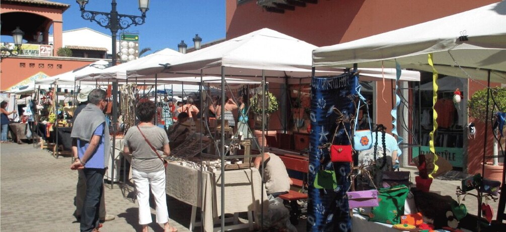 A bustling outdoor market with stalls under white canopies, surrounded by buildings and a clear sky. 3