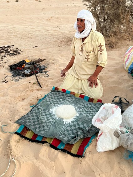 A person kneeling on a sandy beach with a blanket and a ball of dough. 4