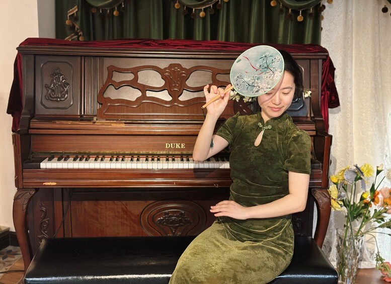 A person sitting on a bench in front of a Duke piano, holding a fan. 1