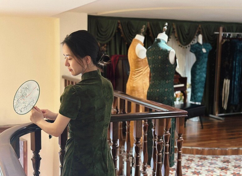 A person in a green dress holding a mirror, standing in a room with a staircase and mannequins displaying dresses. 2