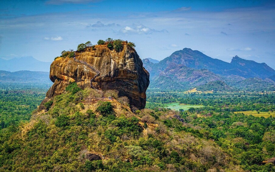 A prominent rock formation with a staircase leading to its summit, surrounded by lush greenery and distant mountains. 3