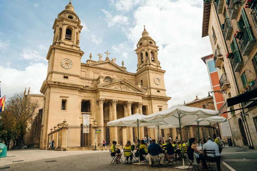 A historic church with two bell towers and a clock, surrounded by a cobblestone square with outdoor seating. 3