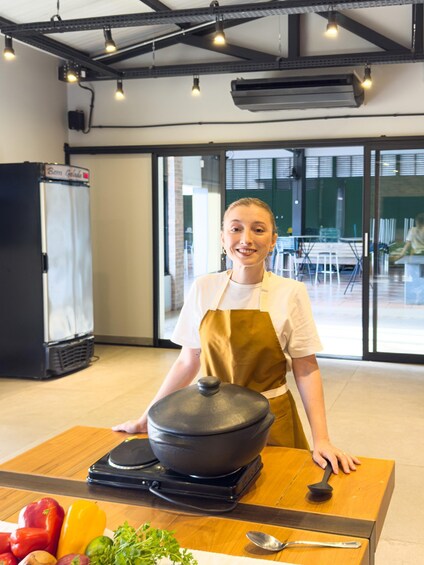 A person in an apron standing behind a table with a pot on a portable stove, surrounded by vegetables. 4