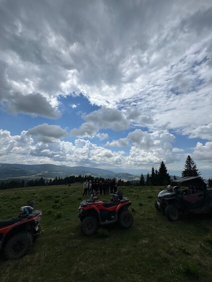 A group of people standing on a grassy hill with mountains in the background and three ATVs parked nearby. 4
