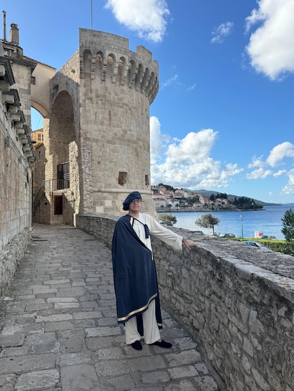 En person klädd i historisk dräkt står på en stenlagd gångstig intill en slottsmur och blickar ut över en kuststad och havet. 2