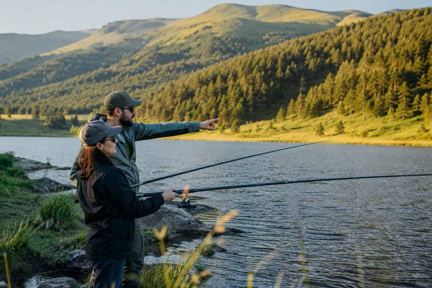 Two people fishing by a calm lake with mountains in the background. 1