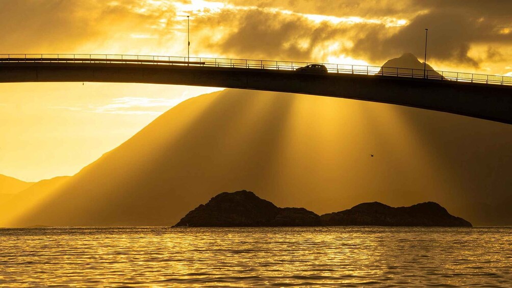 A bridge over water with mountains in the background and sunlight streaming through the clouds. 1