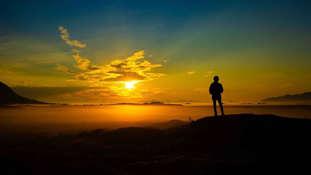 A person standing on a hill overlooking a sunset over a body of water with mountains in the distance. 5