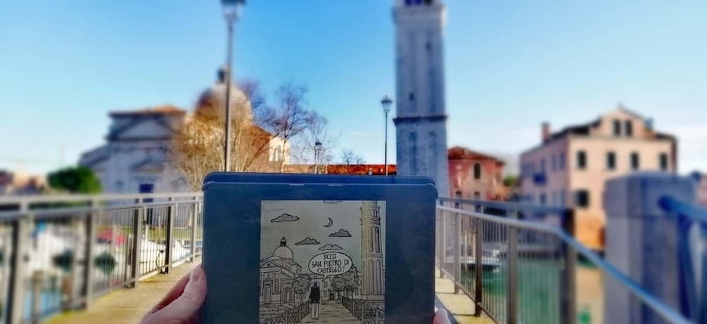A hand holding a book with a drawing of a cityscape, a bridge, and a tower in the background. 1