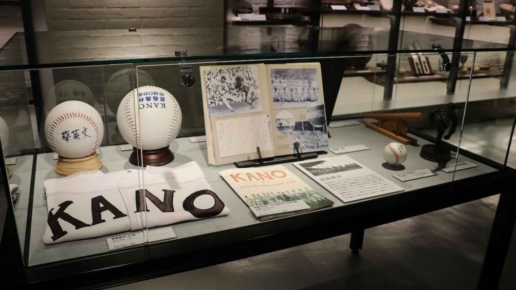 A display case with baseballs, a book titled 'KANO,' and some documents. 2