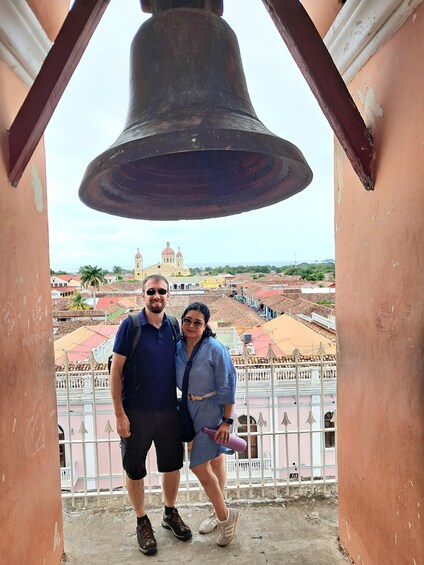 A couple standing in front of a bell, overlooking a cityscape with historic buildings and a church. 3