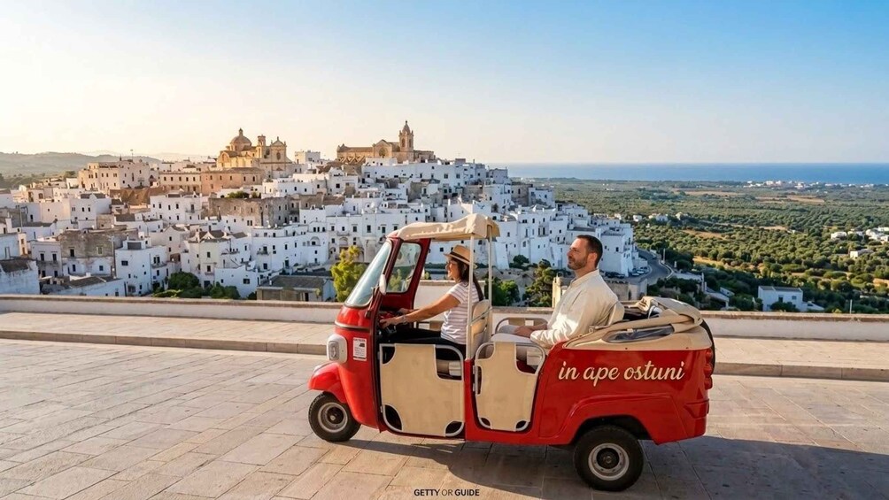 A red three-wheeled vehicle with two passengers, a man and a woman, driving on a paved road with a view of a white-washed town and a church in the background. 1