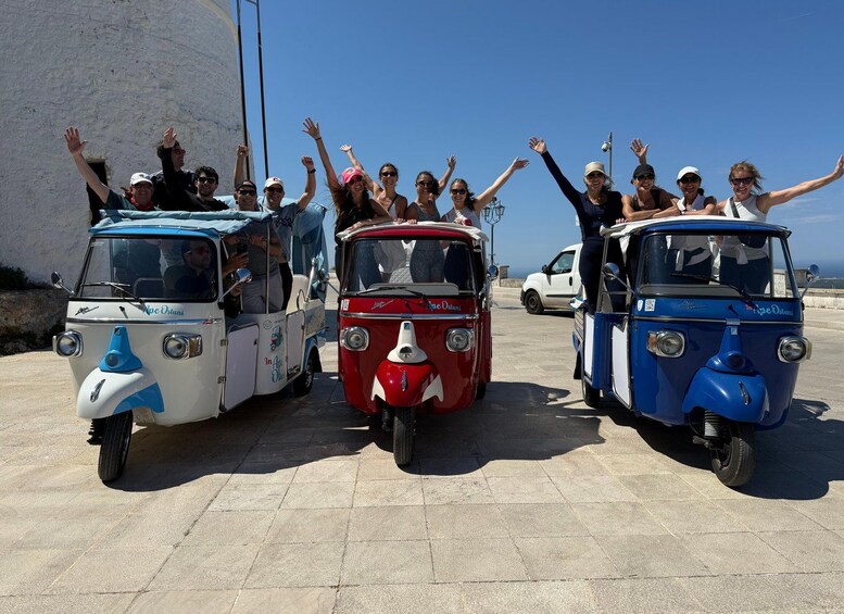 Three vintage cars with passengers raising their hands, parked on a paved surface with a clear sky in the background. 4