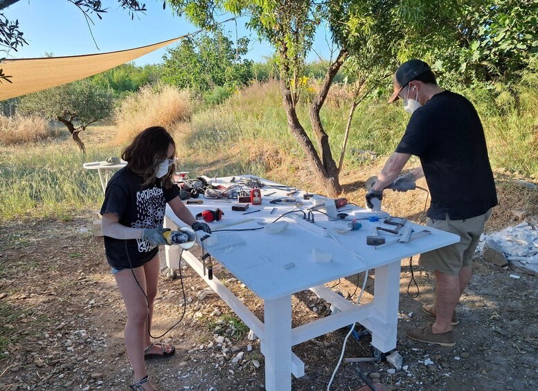 Two people working outdoors at a table with tools and materials, surrounded by trees and dry grass. 3