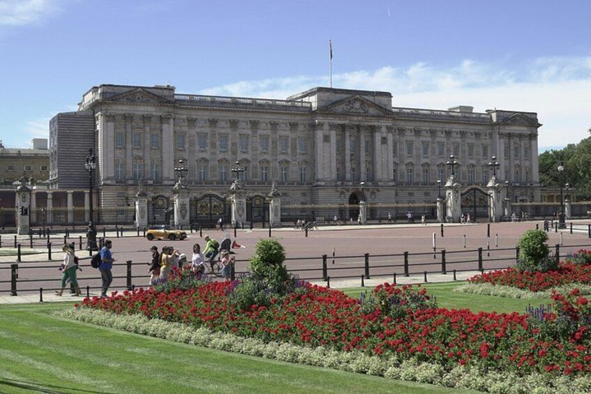 Buckingham Palace, a large historic building with a formal entrance, surrounded by a well-maintained garden with red flowers. 1