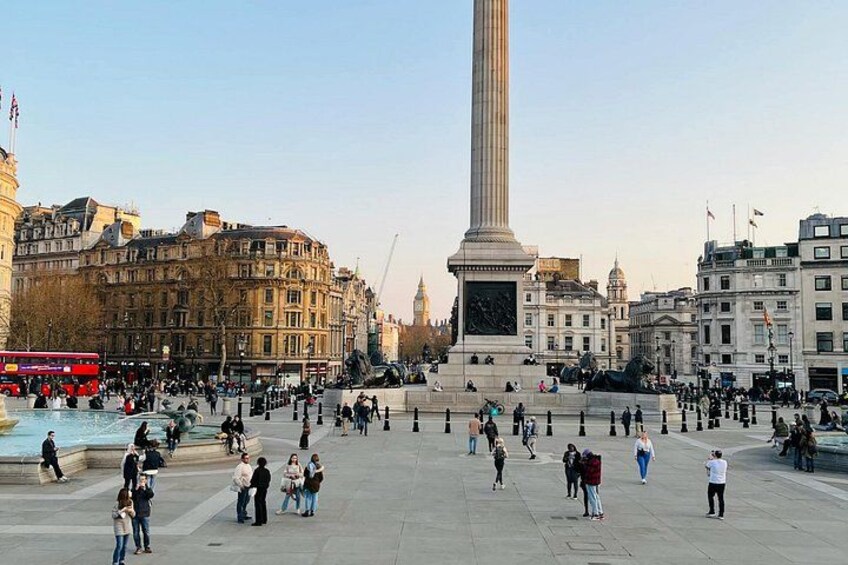 A large open square with a prominent monument, a red double-decker bus, and historic buildings. 3