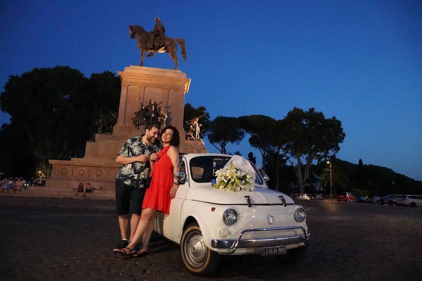 A couple posing in front of a monument with a statue of a horse and rider, a white vintage car, and a statue of a man in a toga. 1
