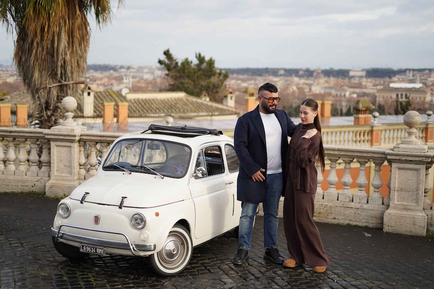 A couple standing beside a vintage white car on a cobblestone path with a cityscape in the background. 3