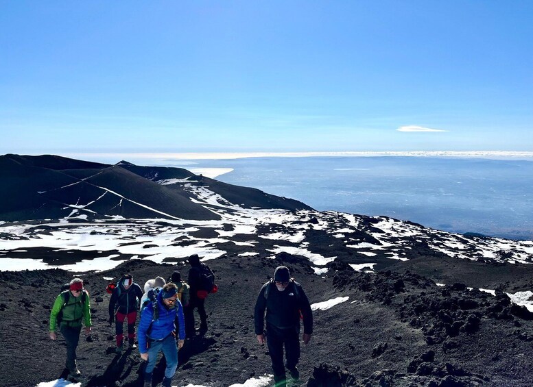 A group of hikers on a volcanic terrain with snow patches and a distant view of a coastline. 1