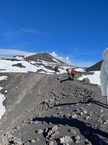 A group of hikers on a rocky mountain trail with snow patches and a volcano in the background. 4
