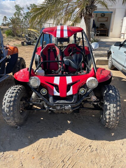 A red off-road vehicle with a license plate, parked on sandy ground with palm trees and a building in the background. 3