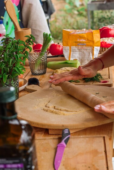 A person rolling dough on a wooden board with a rolling pin, surrounded by kitchen items like a plant, a glass, and a bag of flour. 1