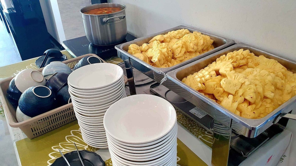 A buffet with various food items, including a pot of soup, trays of pineapple chunks, and stacks of white plates. 2