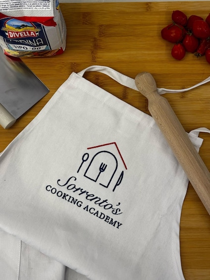 A white apron with 'Sorento's Cooking Academy' embroidered on it, a wooden rolling pin, cherry tomatoes, a metal spatula, and a box of 'DIVELLA' pasta on a wooden surface. 3