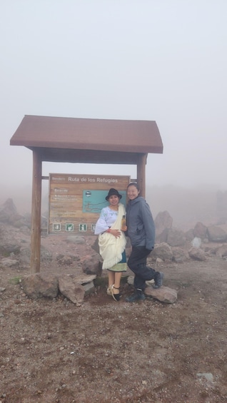 Two people standing in front of a wooden signpost with information about Sendero Ruta de los Refugios. 1