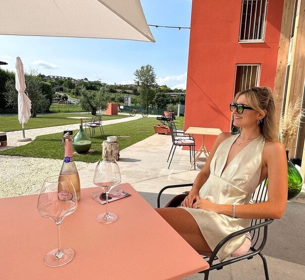 A person seated at an outdoor table with wine glasses and a bottle. 1
