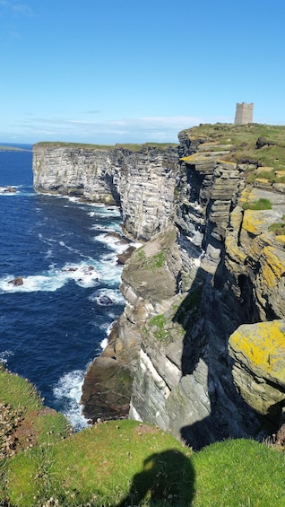 A coastal cliff with waves crashing against it, a grassy area, and a tower in the distance. 2