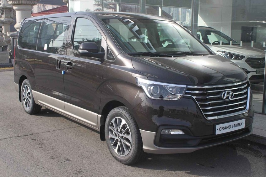 A dark-colored Hyundai Grand Starex van parked in front of a building with large windows. 3
