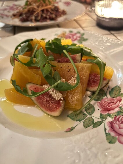 A plate of food with figs, orange slices, and greens, on a floral tablecloth. 3