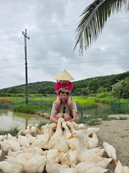 A person with a child on their shoulders, surrounded by white ducks, with a water body and greenery in the background. 4