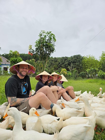 Three people sitting on the grass surrounded by a flock of white ducks. 5