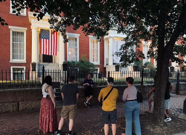 A group of people standing on a brick sidewalk, facing a building with a large American flag. 3