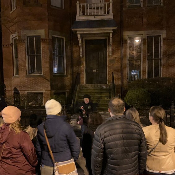 A group of people standing in front of a brick building with a dark entrance and a small porch. 2