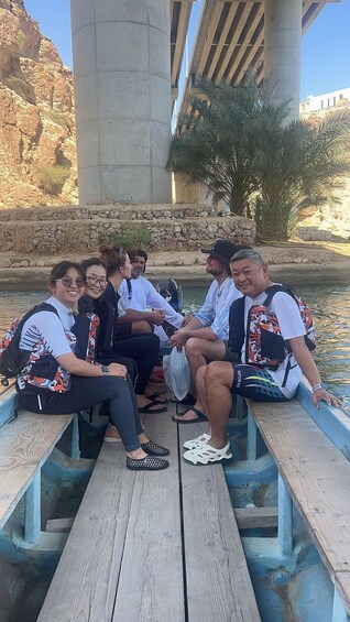 A group of people sitting on a wooden dock under a bridge, with a rocky landscape and palm trees in the background. 5