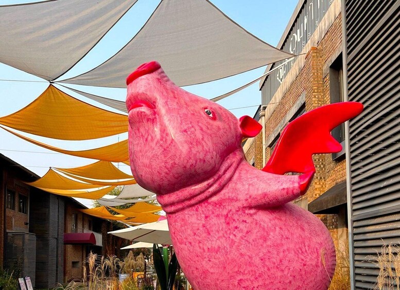A pink pig sculpture with red ears and a red snout, set against a backdrop of buildings and yellow and white umbrellas. 4