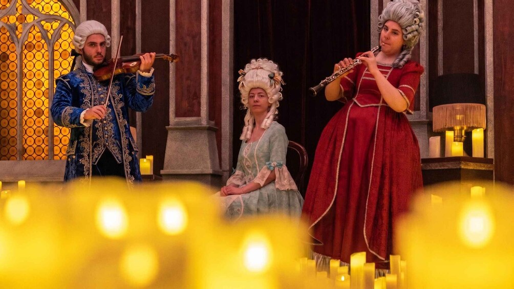 Three people in period costumes playing musical instruments on a stage with candles. 1