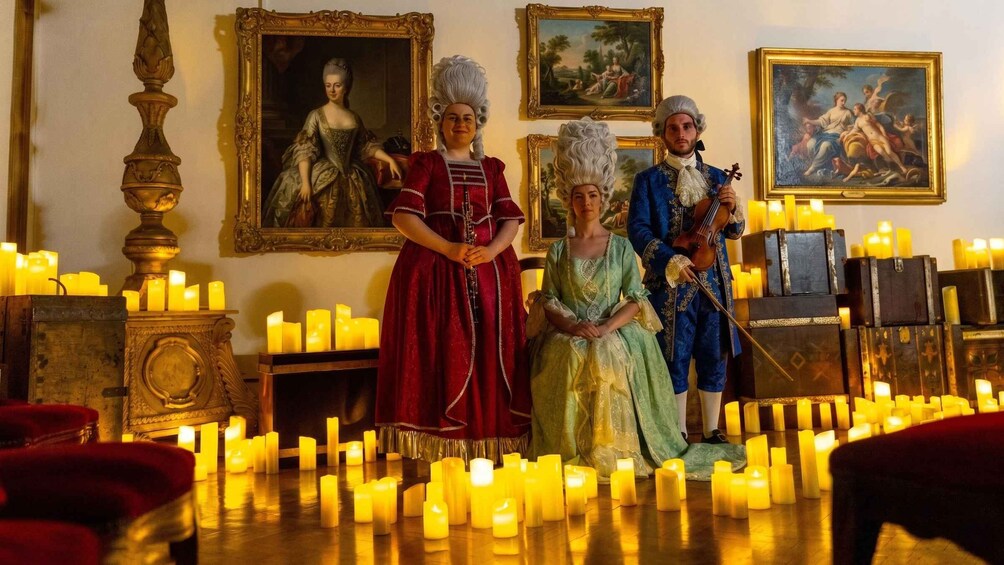 Three people in period costumes, a room with classical paintings, and numerous lit candles. 3