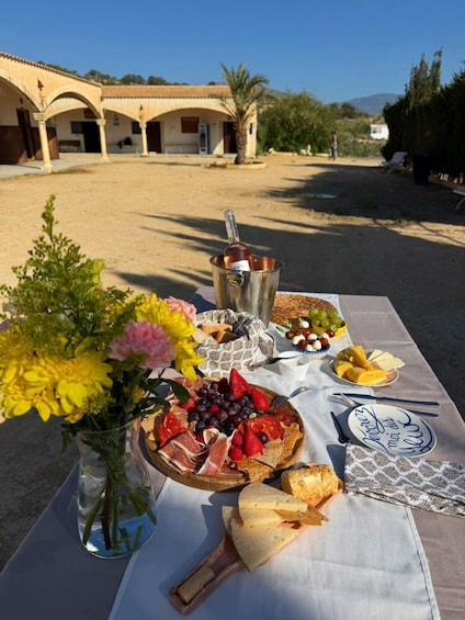 An outdoor dining setup with a table set for a meal, including a variety of cheeses, fruits, and bread, with a bottle of wine in the background. 4