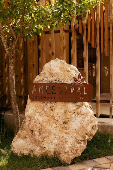 A large rock with a sign that reads 'ANCESTRAL CULINARY EXPERIENCE' in front of a wooden fence and a tree. 2
