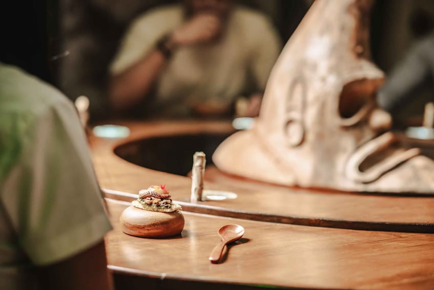 A wooden table with a carved wooden sculpture, a small bowl with food, and a spoon. 1
